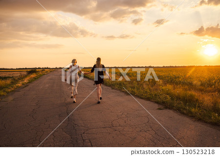 Two women walking hand in hand along a country road at sunset near blooming sunflower fields 130523218