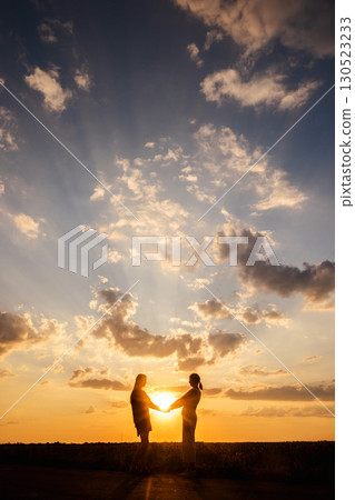 Silhouette of two women standing in a field and holding hands during a colorful sunset with dramatic rays of light in the sky 130523233