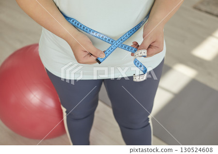 Overweight woman in sportswear is holding centimeter tape and measuring her waist 130524608