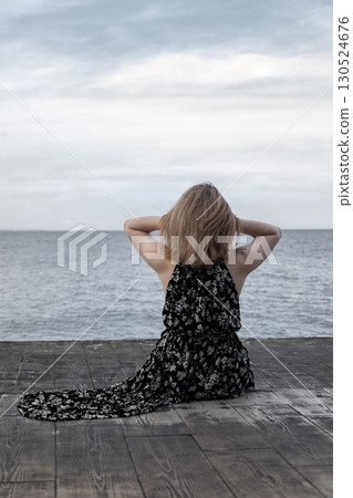 Woman in dress sitting on wooden pier by the sea, back view. Concept of relaxation, solitude, freedom, summer vacation and peaceful lifestyle Woman in dress sitting on wooden pier by the sea, back view. Concept of relaxation, solitude, freedom, summer vacation and peaceful lifestyle 130524676