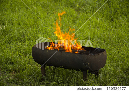 Bright fire burning in barbecue grill on green grass. Concept of outdoor cooking, picnic preparation, camping, summer lifestyle and fire element Bright fire burning in barbecue grill on green grass. Concept of outdoor cooking, picnic preparation, camping, summer lifestyle and fire element 130524677
