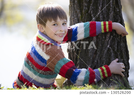 A little boy in a knitted sweater sits near a tree and looks at the camera. 130526200