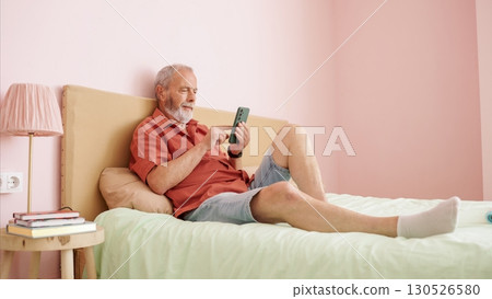 Senior man relaxing on bed using smartphone in modern apartment 130526580