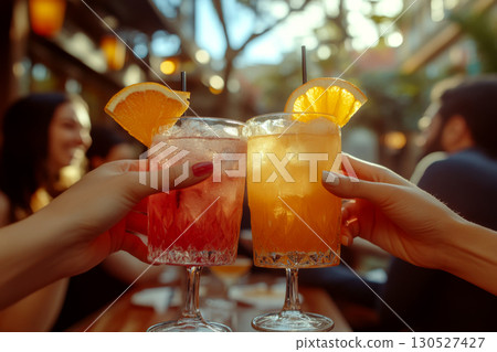 Two hands hold colorful cocktails with orange slices in an outdoor setting. Concept of social gathering. For beverage advertisement. 130527427