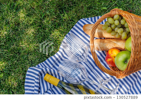 Picnic basket with wine, fruits, and baguette on a sunny green meadow 130527889