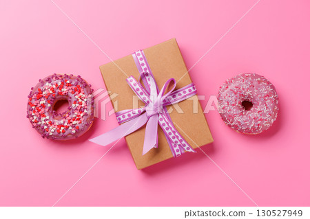 A festive flat lay featuring vibrant donuts with colorful toppings and a gift box on a pink background 130527949