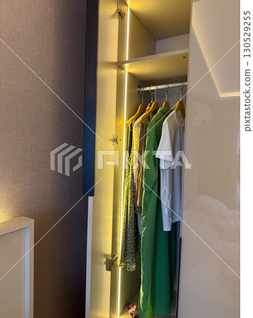 Wardrobe with hanging clothes including shirts and dresses on wooden hangers. Interior design, fashion lifestyle, and daily organization in modern living space. Wardrobe with hanging clothes including shirts and dresses on wooden hangers. Interior design, fashion lifestyle, and daily organization in modern living space. 130529255