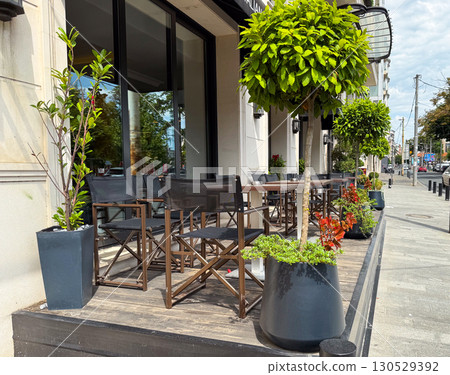 Cafe terrace with arranged tables, chairs, and potted trees along city sidewalk. Outdoor dining, urban lifestyle, and hospitality design in commercial restaurant setting. 130529392