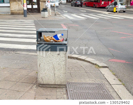 Public trash bin with plastic bottle and paper waste near crosswalk in urban street. Environmental concern, waste disposal, and sanitation issue in modern city life. 130529402