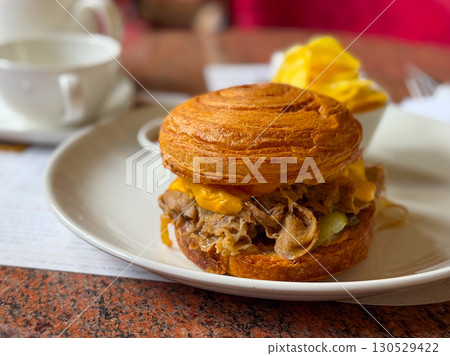 Croissant sandwich with meat and melted cheese served on a plate in a cozy cafe. Gastronomy, lifestyle, and casual dining experience with comfort and indulgence. Croissant sandwich with meat and melted cheese served on a plate in a cozy cafe. Gastronomy, lifestyle, and casual dining experience with comfort and indulgence. 130529422