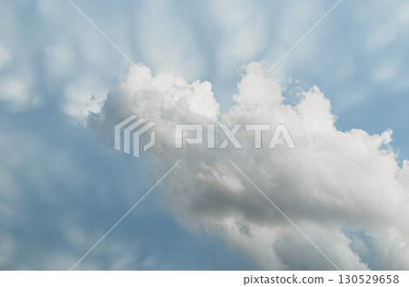 billowing white cumulus cloud drifts across a soft blue sky, with dappled light casting gentle 130529658