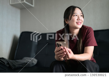 Young Thai woman sitting on sofa using smartphone at home, lifestyle portrait 130530021