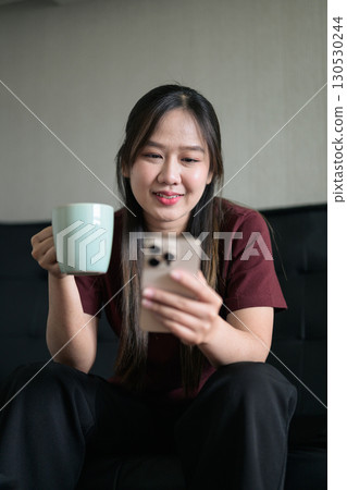 Young Thai woman sitting on sofa using smartphone at home and drinking coffee Young Thai woman sitting on sofa using smartphone at home and drinking coffee 130530244