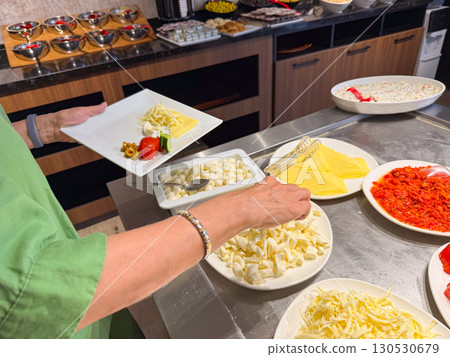 Person selecting cheese and vegetables at buffet with plate in hand. Lifestyle, hospitality, and nutrition in hotel breakfast environment. Person selecting cheese and vegetables at buffet with plate in hand. Lifestyle, hospitality, and nutrition in hotel breakfast environment. 130530679