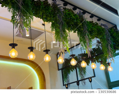 Row of glowing bulbs hanging under green foliage decoration indoors. Lighting, hospitality design, and lifestyle atmosphere in modern interior. 130530686