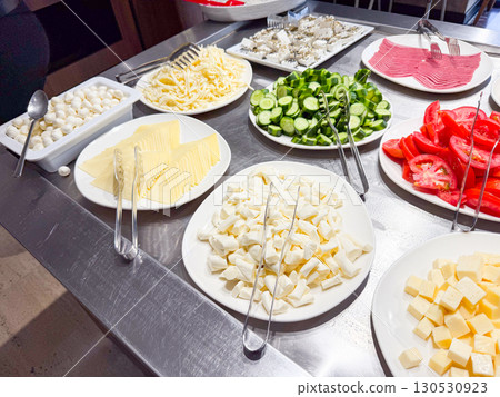 Plates with sliced cheese, vegetables, and cold cuts arranged on breakfast buffet table. Food, nutrition, and hospitality service in hotel dining area. 130530923