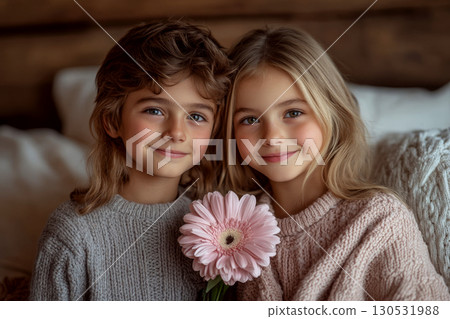 Two children sit close together indoors, holding a pink flower and smiling. Concept of friendship and joy. For family-themed content. 130531988
