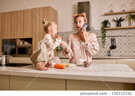 Mother multitasking on phone while bonding with daughter in modern kitchen 130532292