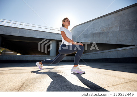 Young woman performing fitness lunges outdoors in a sunny urban environment Young woman performing fitness lunges outdoors in a sunny urban environment 130532449