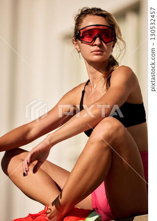Athletic woman wearing red-tinted glasses taking a break in outdoor sunlight 130532457