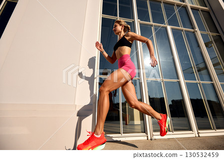 Athletic woman running outdoors next to modern glass building in bright sunlight 130532459
