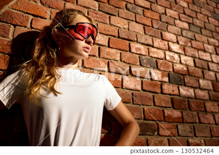 Young woman wearing sports sunglasses standing by a brick wall in sunlight Young woman wearing sports sunglasses standing by a brick wall in sunlight 130532464