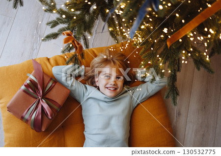 Smiling child top view under Christmas tree with gift box on orange cushion on wooden floor Smiling child top view under Christmas tree with gift box on orange cushion on wooden floor 130532775