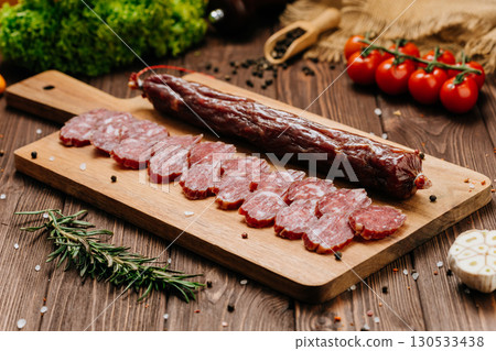 Sliced dry sausage on rustic wooden board with spices and tomatoes, top view 130533438