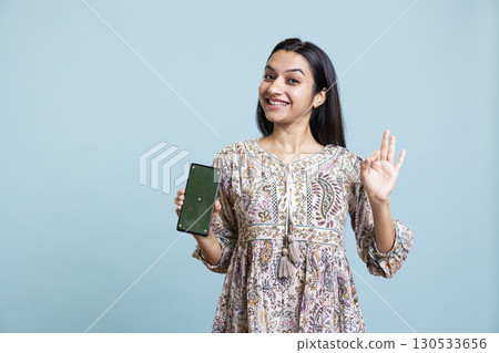 Portrait of indian woman doing marketing ad with green screen device, standing against blue background. Smiling young adult feeling positive on an advertisement in front of the camera. 130533656