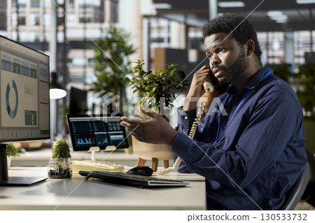 African american worker engages in a landline phone call at the office, managing a project briefing and discussing tasks completion with the supervisor. Multitasking for productivity. 130533732