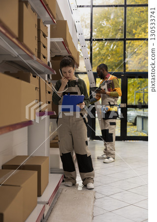 Staff member answering a call in small scale logistics facility, confirming online orders and handling shipment service operations. Small business delivery preparation management. Staff member answering a call in small scale logistics facility, confirming online orders and handling shipment service operations. Small business delivery preparation management. 130533741