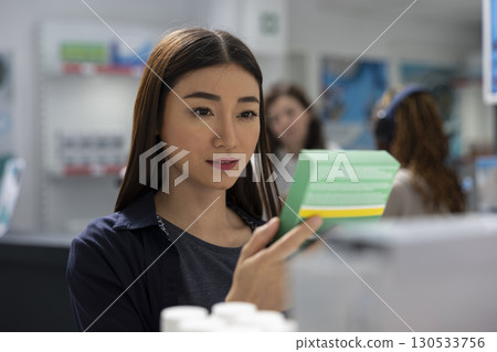 Customer reading labels information on vitamins placed on pharmacy shelves, retail service in a healthcare space. Asian woman searching for wellness supplements, pharmacy visit. 130533756