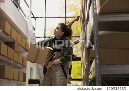 Woman focused on in house shipment operations carrying cardboard boxes for dispatch and ensuring e-store logistics. Staff processing orders and preparing parcels, meeting retail standards. 130533885