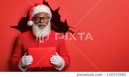 African American Santa, Black Santa Claus with red box his hands looks through torn hole red paper background jumping, points his hand empty place copy space background. 130535060