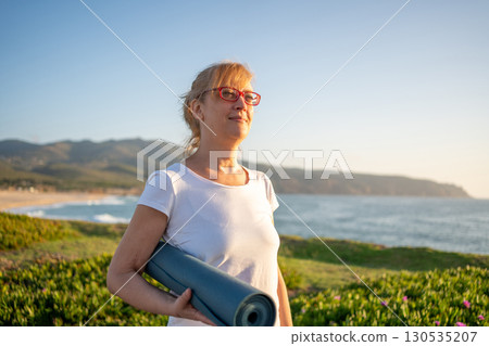 Pensioner woman holding yoga mat standing on ocean Pensioner woman holding yoga mat standing on ocean 130535207