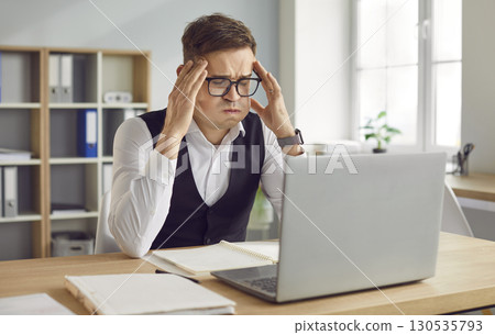 Tired young exhausted overworked business man sitting at the desk on his workplace in office. Tired young exhausted overworked business man sitting at the desk on his workplace in office. 130535793