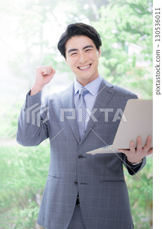 A man in a suit holding a laptop in the soft greenery of the backlight looks at the camera and gives a fist pump. A man in a suit holding a laptop in the soft greenery of the backlight looks at the camera and gives a fist pump. 130536011