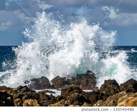 Powerful Ocean Wave Crashing Against Dark Rocks Creating White Foam and Spray Under a Cloudy Blue Sky 130540490