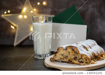 Christmas Stollen with glass of milk for SANTA and envelope with letter in Christmas lights and decoration. Traditional sweet fruit loaf with icing sugar festive pastry dessert. Holiday baking food 130541241