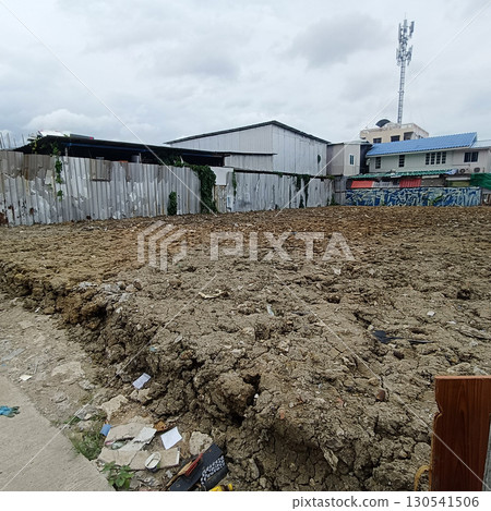 Vacant plot of land with dry, cracked soil and scattered debris, surrounded by metal fence and neighboring buildings under cloudy sky, conveying urban environment and construction potential 130541506