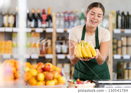 Young woman seller offering bananas in grocery store Young woman seller offering bananas in grocery store 130541784