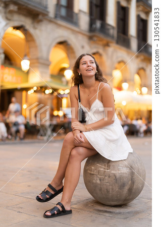 Evening Royal square in Barcelona, girl in sundress is sitting on stone ball 130541833