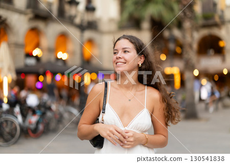 In evening, young smiling girl walks along city street Placa Real 130541838