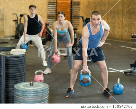 Active middle-aged man in sportswear exercising CrossFit with kettlebell while training with group in fitness center Active middle-aged man in sportswear exercising CrossFit with kettlebell while training with group in fitness center 130542702