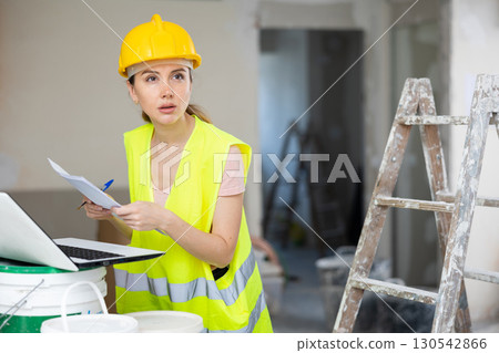 Female foreman in a protective helmet and a yellow vest checks the execution of repair work using laptop 130542866
