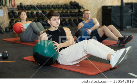 Strong young man performing exercise for abs core with medicine ball on yoga mat during group workout in CrossFit gym center 130542919