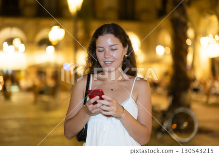At night, smiling girl with smartphone walks along city street Placa Real 130543215