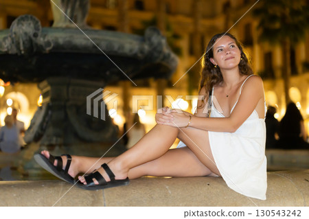 Night Royal square in Barcelona, girl in sundress is sitting on fountain fencing. 130543242
