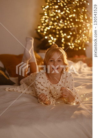 Smiling girl in white dress lying on bed with Christmas tree lights in cozy room Smiling girl in white dress lying on bed with Christmas tree lights in cozy room 130543529