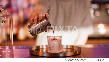 Pouring cocktail. Cold aperitif. Male barman adding red liquid from metal shaker to glass. Making big ice cube negroni on pub counter. Pouring cocktail. Cold aperitif. Male barman adding red liquid from metal shaker to glass. Making big ice cube negroni on pub counter. 130543642